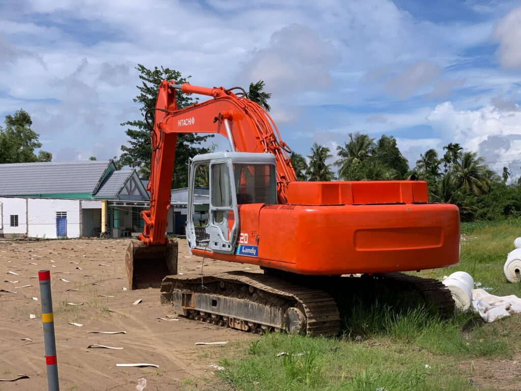 An orange excavator stands idle at a sunny construction site with tropical greenery.