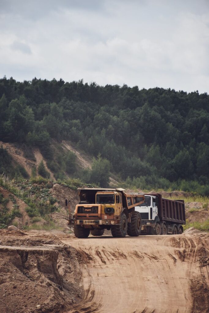 Two heavy-duty trucks on a dirt road in a forest quarry with a lush backdrop.